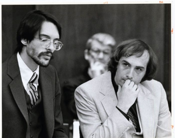 Imagem de Billy Milligan durante seu julgamento: à esquerda, aparentemente, seu advogado, homem magro de óculos e barba vestindo terno preto. à direita Milligan com mão no queixo e vestindo blazer claro