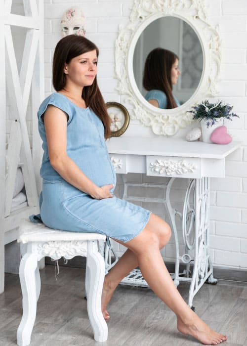 mulher gravida sentada ao lado de penteadeira com a mao sobre o ventre Queda de cabelo na gravidez: causas, tratamento e dicas para lidar com esse "probleminha".